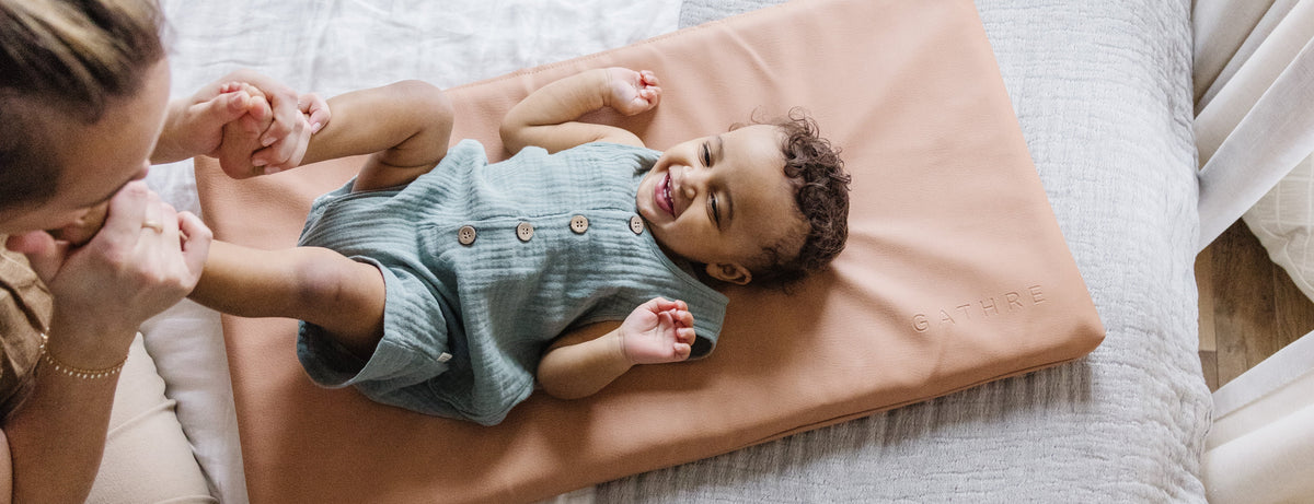 A baby in a blue-green romper lies on a peach-colored changing pad on a bed, while a woman holds the baby's feet. The scene is shot from an overhead angle in a bright, neutral-toned room.
