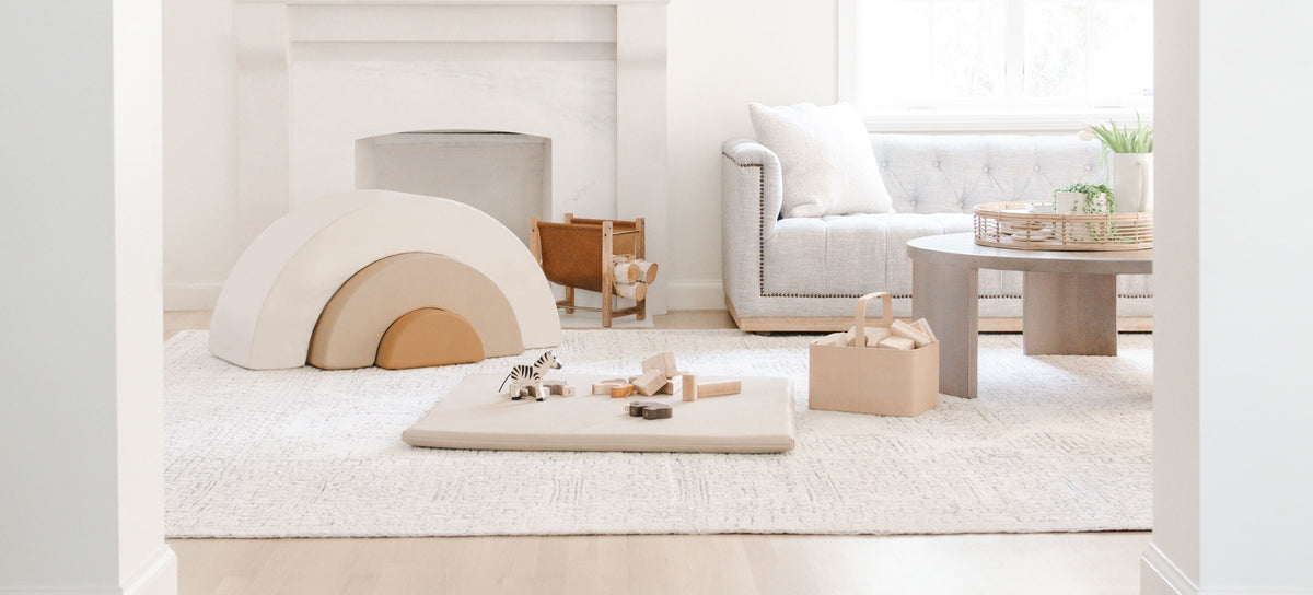 A bright, modern living room viewed through a large white arched opening with light wood steps in the foreground. The room features a white fireplace, a light gray sofa, a cream rug, and a minimalist coffee table.