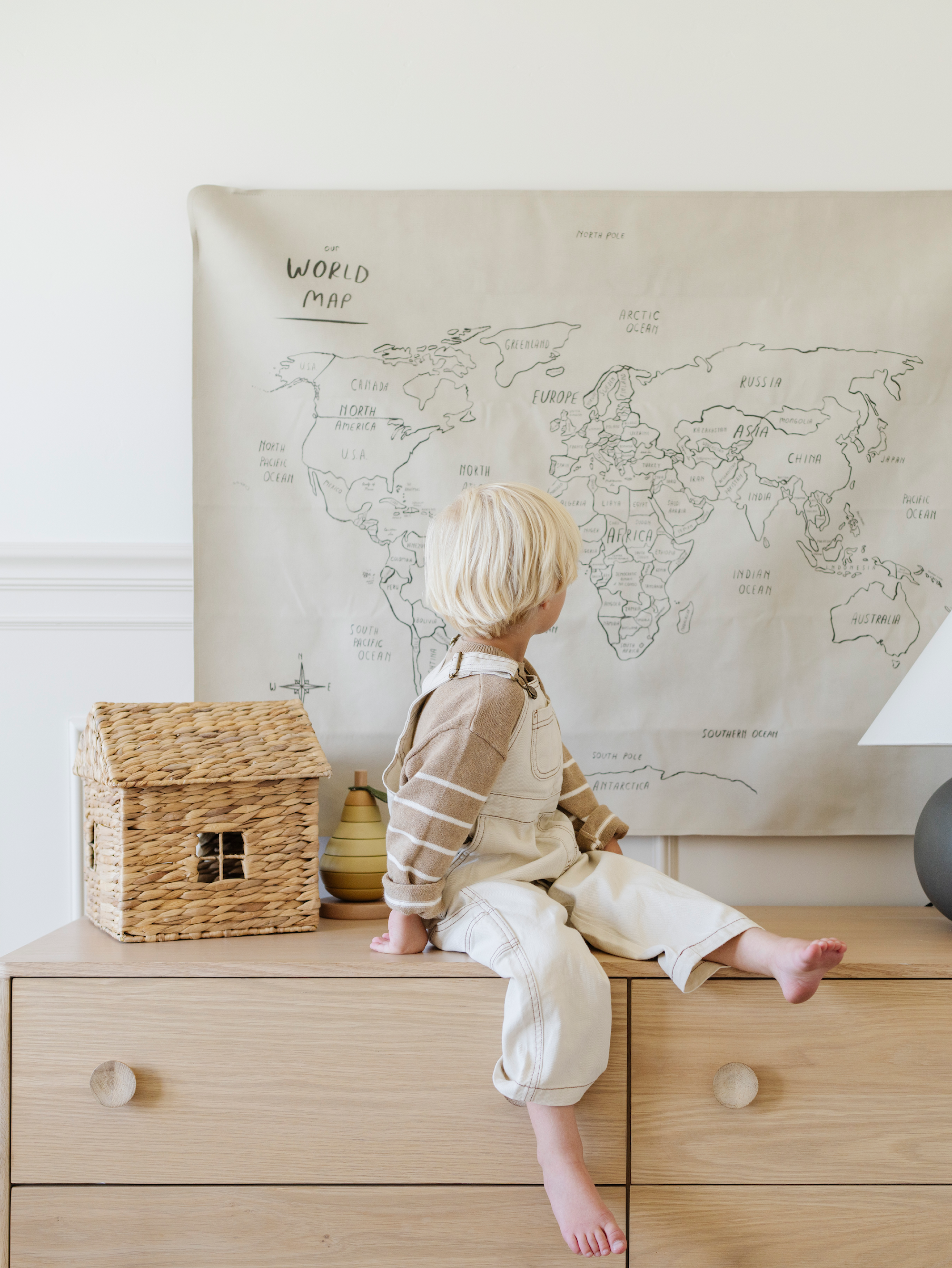 Little kid sitting by a Gathre Tapestry hanging on a wall.